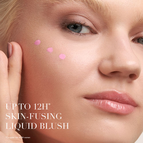 A close-up of a young woman applying liquid blush to her cheek, showcasing a smooth complexion and three small dots of pink blush. The text highlights "UP TO 12H SKIN-FUSING LIQUID BLUSH."