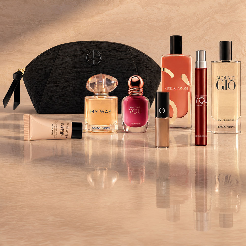Black Armani Beauty pouch displayed with assorted fragrances, makeup and skincare products against a beige backdrop.  
