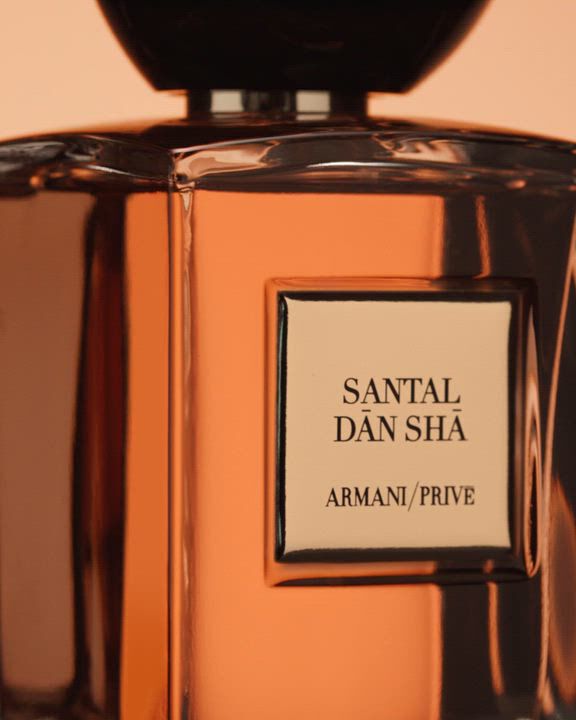 A close-up of an amber-toned "Armani/Prive Santal Dān Shā" fragrance bottle, featuring a dark, rounded cap. The bottle is subtly overlaid with flowing, ethereal brown and orange abstract shapes, and flashes of sparkling red crystal formations.