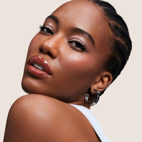 A close-up portrait of a woman with braided hair, showcasing a radiant complexion and glossy lips against a neutral background.
