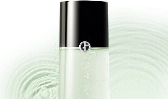 A clear bottle of skincare product with a sleek black cap, featuring a logo on the front, set against a background of soft, rippling green water.
