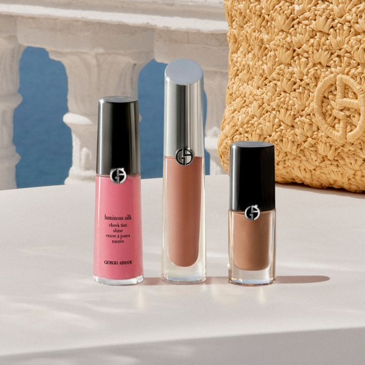 Giorgio Armani makeup products displayed on a white pedestal: a pink liquid blush, a nude lip gloss, and a brown liquid lipstick, set against a Mediterranean-inspired backdrop with stone arches, blue sky and a textured gold accessory.