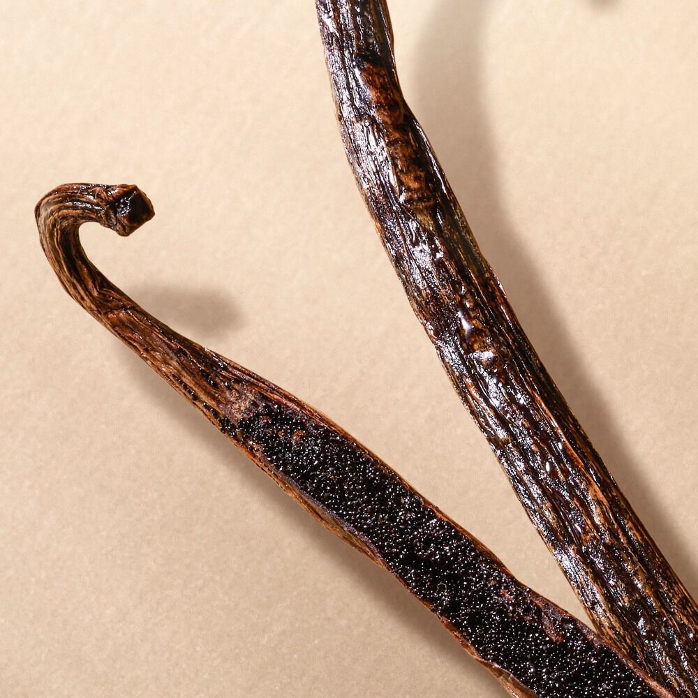 Shiny vanilla bean pods, one split open to reveal seeds, on a beige background.