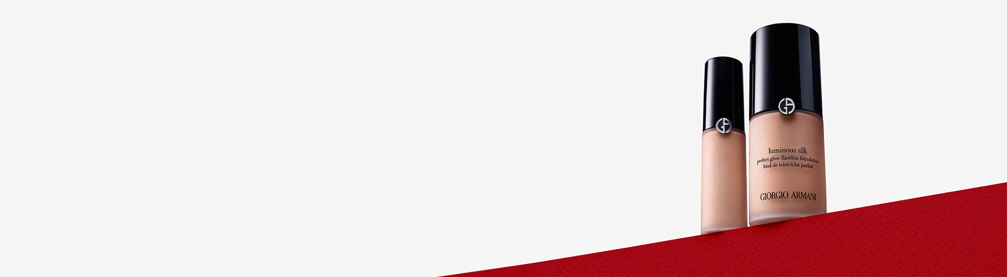 Two bottles of foundation from Giorgio Armani are displayed against a light gray background, with a red section at the bottom. One bottle is larger than the other, both featuring sleek black caps.