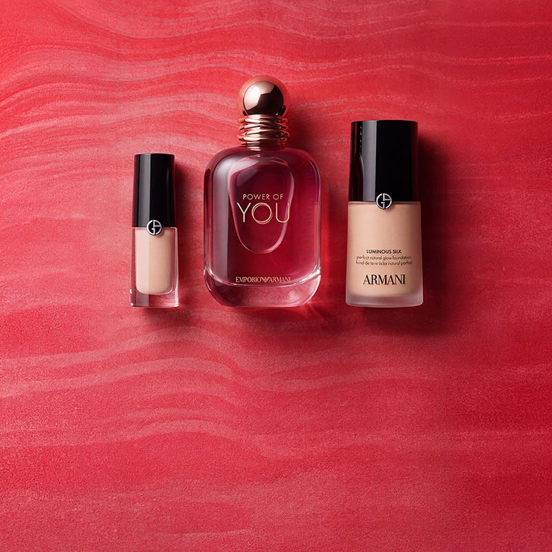 Product flat lay featuring four Armani beauty products arranged on a vibrant red textured background from left to right: the Luminous Silk Foundation in a glass bottle with a black cap and the GA logo, the Luminous Silk Concealer in a slimmer glass bottle, an open Armani lipstick in a metallic silver case, and a liquid highlighter or eye tint in a glass bottle with a black cap.​