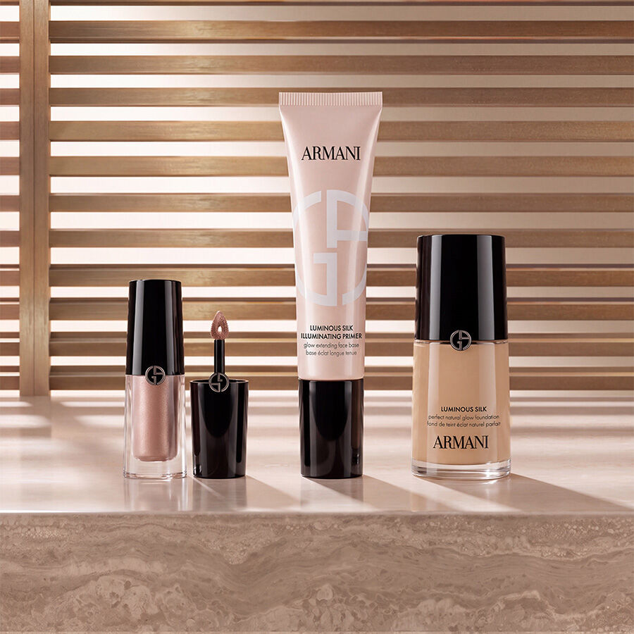 Armani beauty makeup products including liquid highlighter, Luminous Silk Illuminating Primer, and Luminous Silk Foundation displayed on a marble surface against a warm slatted background.