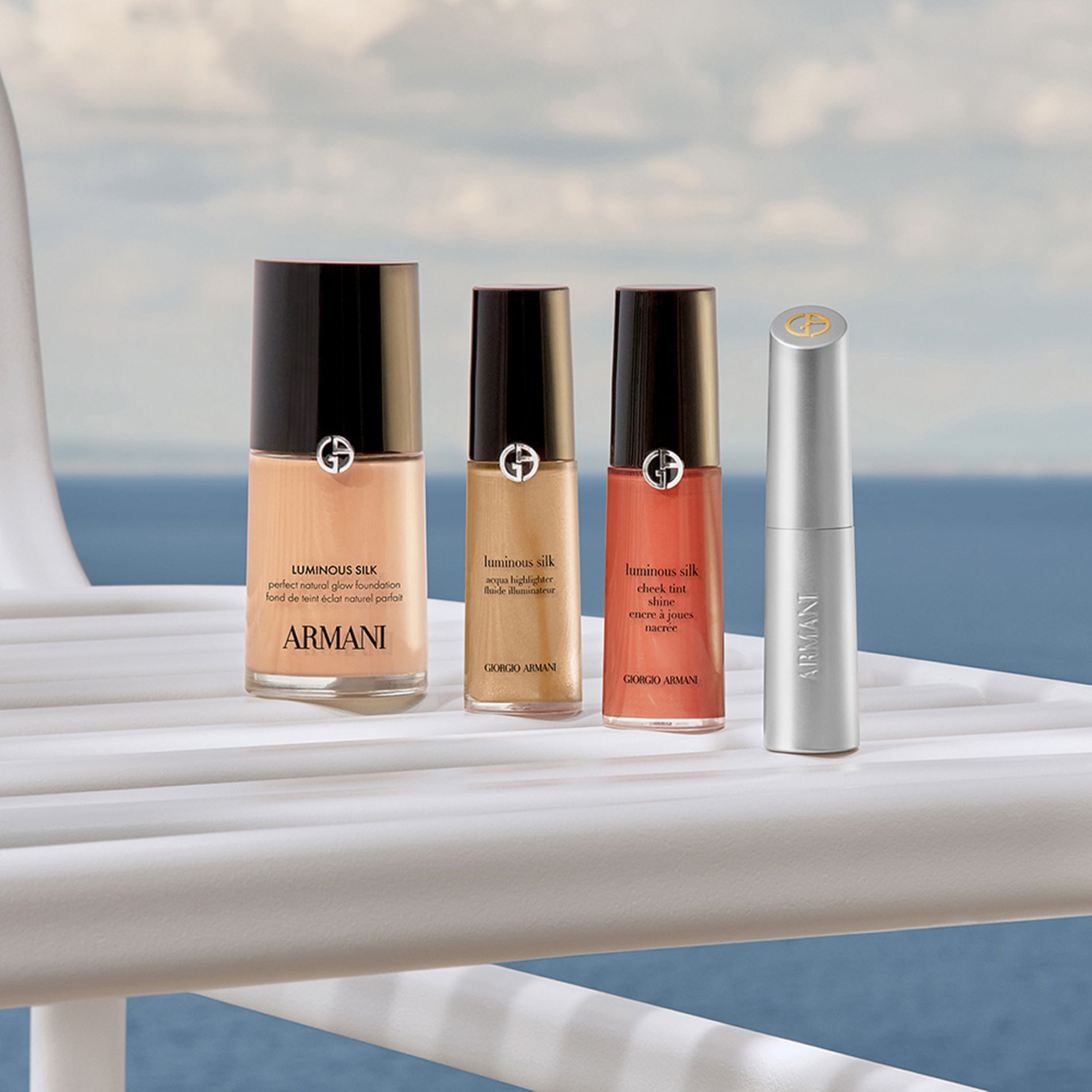 A sophisticated lineup of Armani icons: Luminous Silk Foundation, Concealer, Cheek Tint, and a silver lipstick. Resting on a white yacht railing against the sea, they promise a radiant, effortless look for a luxurious coastal escape.