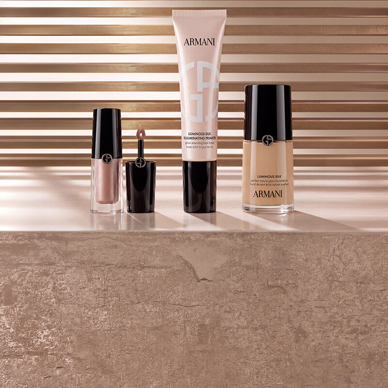 Armani beauty makeup products including liquid highlighter, Luminous Silk Illuminating Primer, and Luminous Silk Foundation displayed on a marble surface against a warm slatted background.