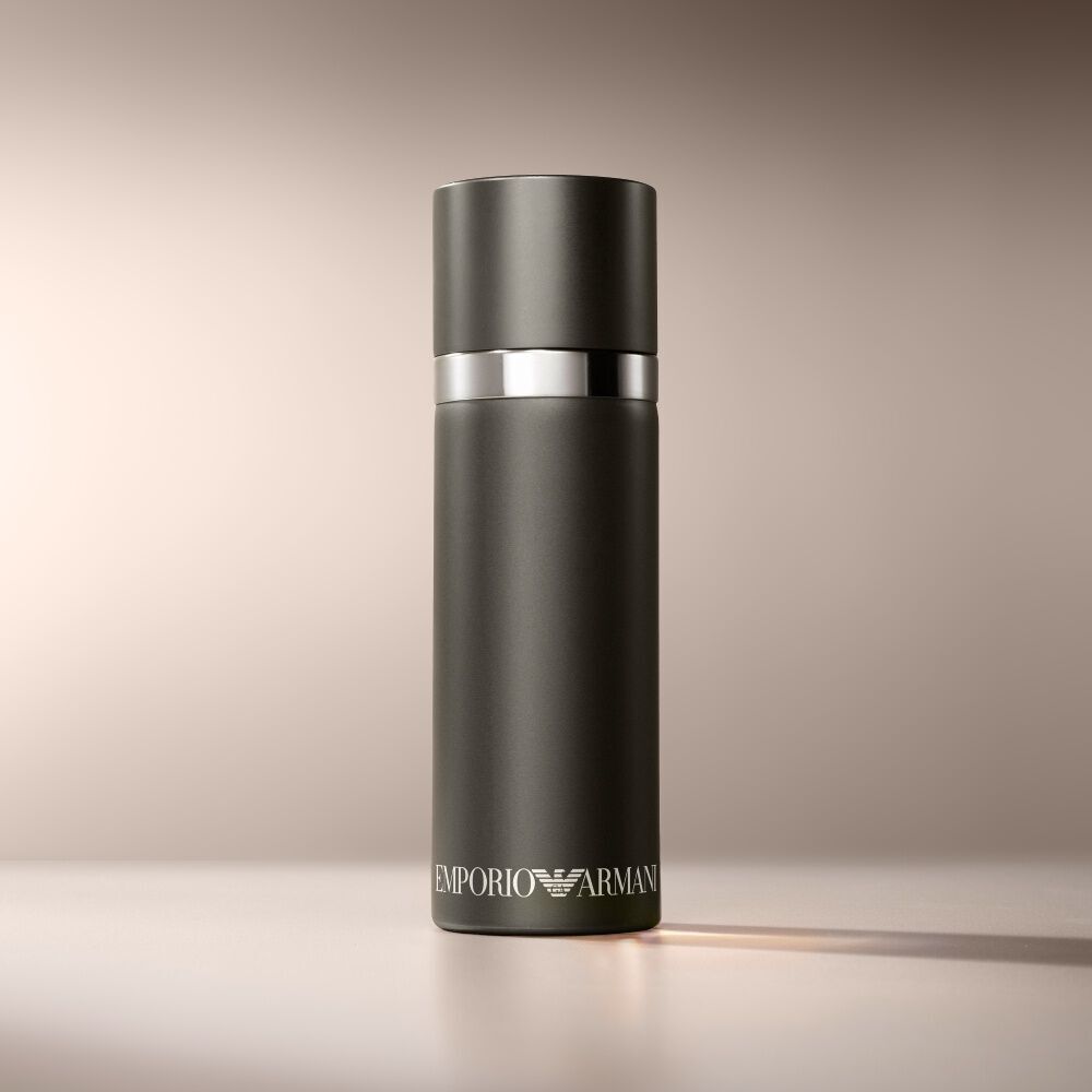 A matte black cylindrical Emporio Armani "He" fragrance bottle is presented on a warm, light brown background in a horizontal aspect ratio.