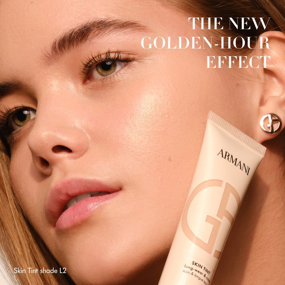 Woman with radiant skin holding an Armani Skin Tint tube, with text THE NEW GOLDEN-HOUR EFFECT.