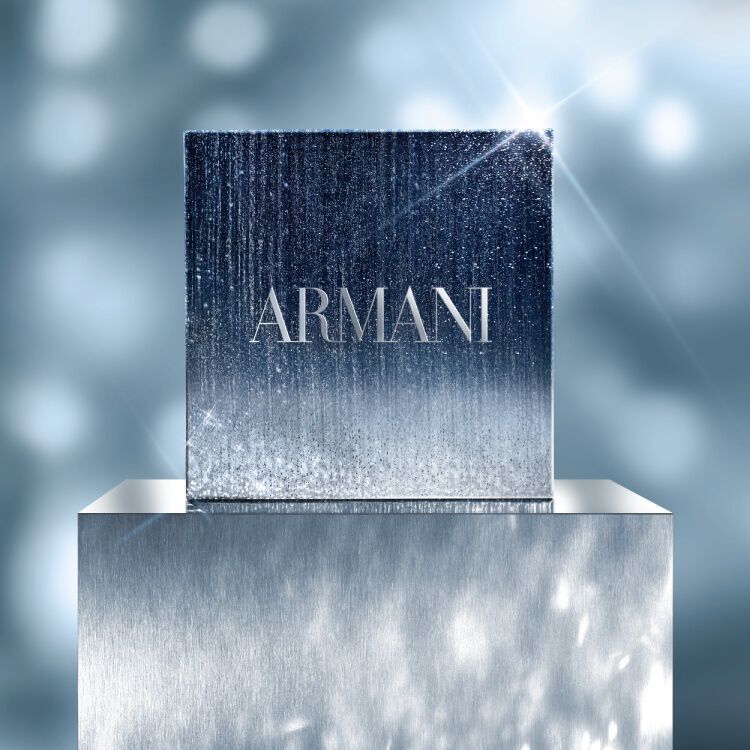 A square Armani GIFT SET in a shimmering silver and blue gradient box, displayed on a reflective pedestal with a soft, sparkling light background.