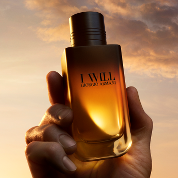 A hand holding the I WILL Giorgio Armani fragrance bottle against a golden sunset sky.