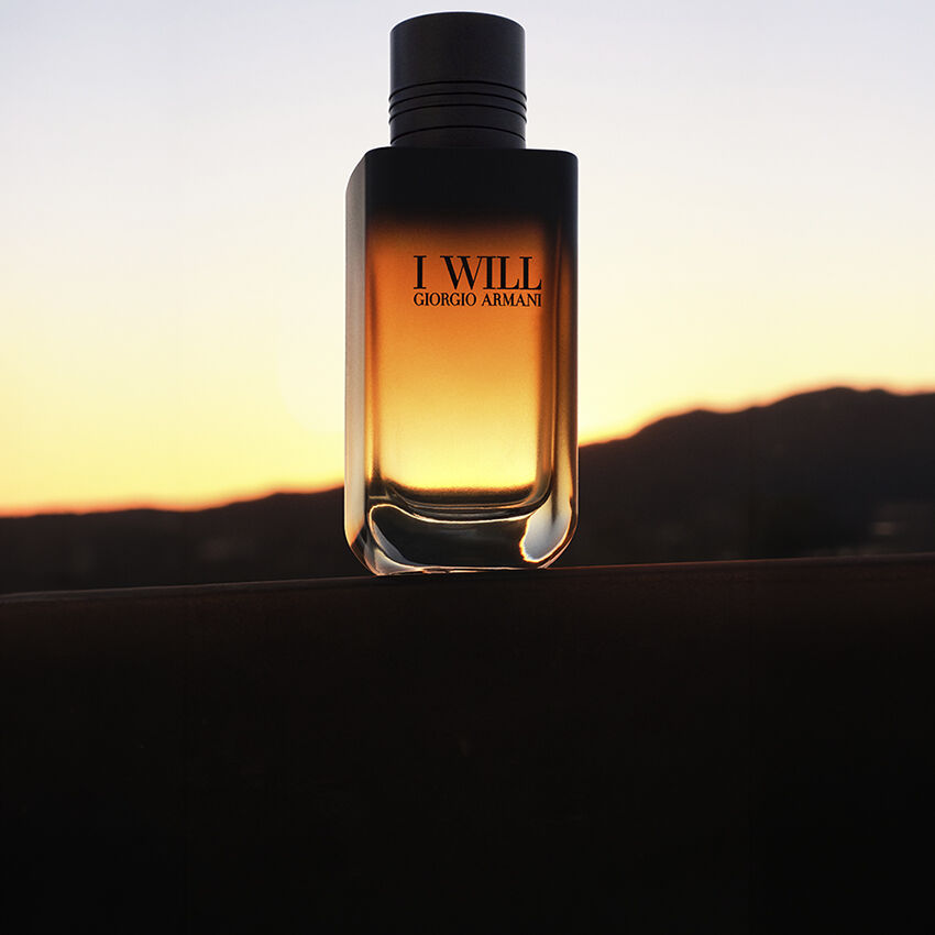 A hand holding the I WILL Giorgio Armani fragrance bottle against a golden sunset sky.