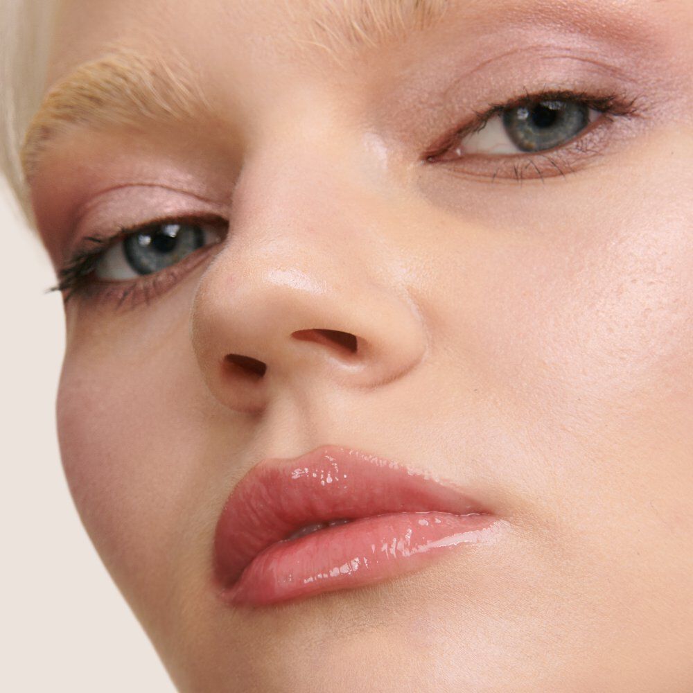 Close-up of a model's face featuring pale skin, light pink eye makeup, and glossy lips, with a soft focus background.
