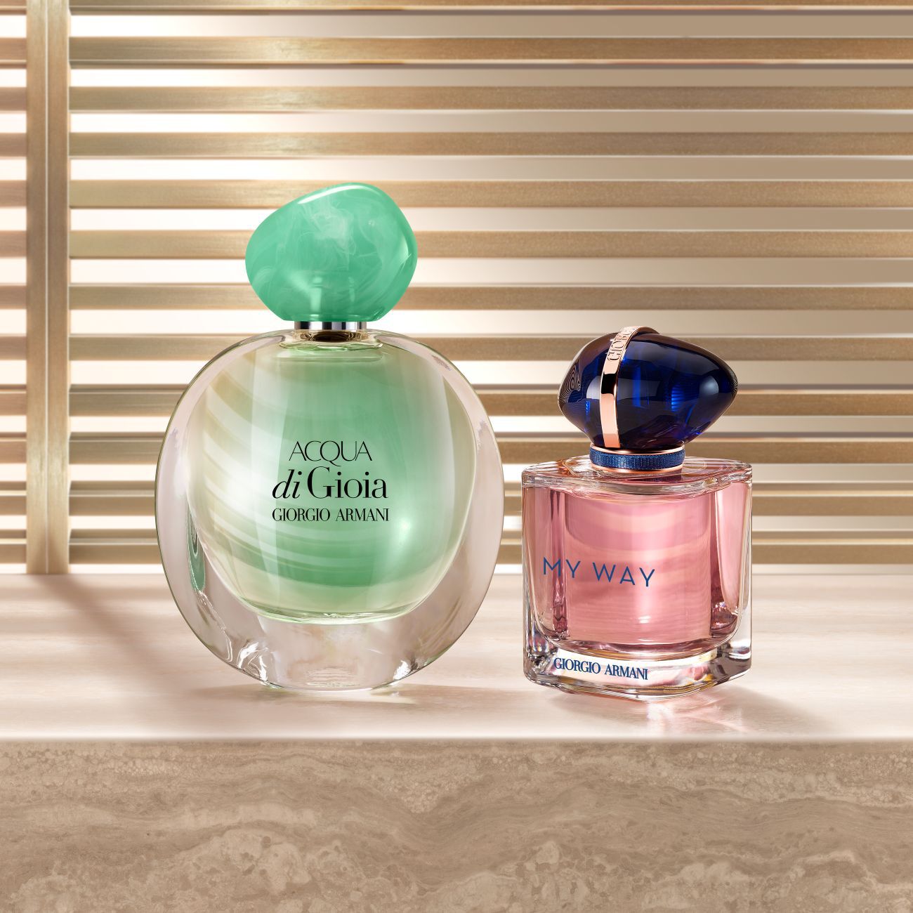 Two Giorgio Armani perfume bottles, Acqua di Gioia in green and My Way in pink, displayed on a stone surface against a gold slatted background.