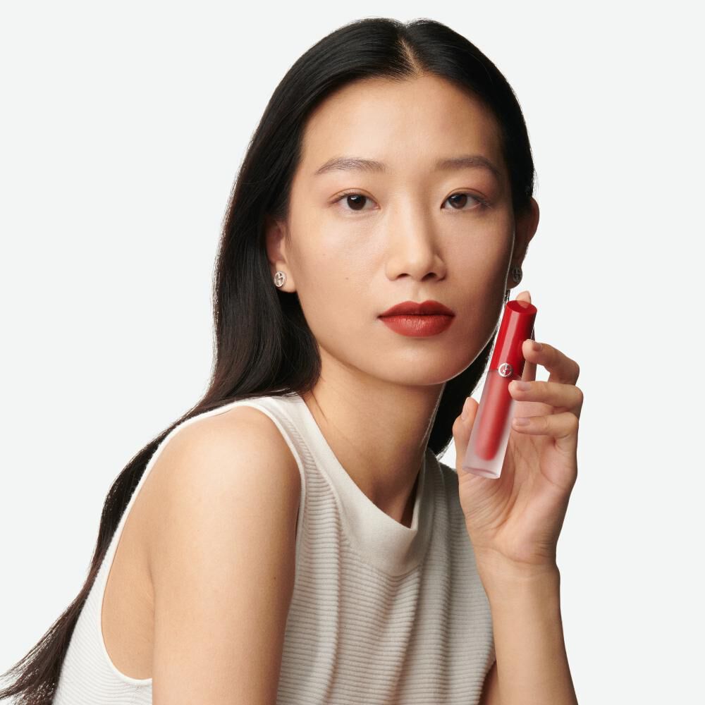 A woman with long black hair holds a red lipstick in her hand, wearing a beige sleeveless top and a subtle makeup look.