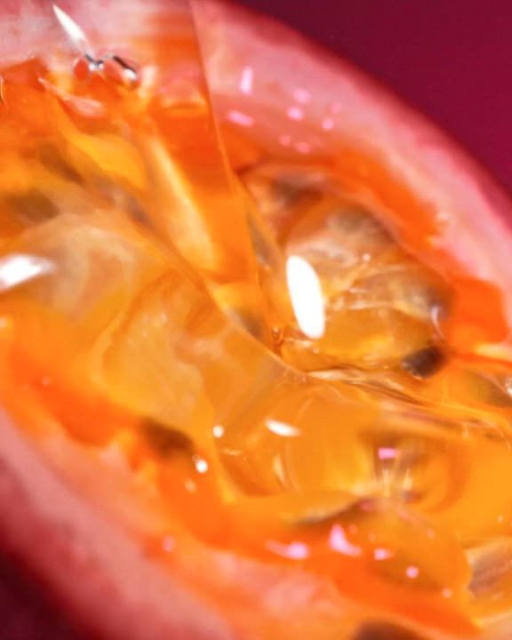 An extreme close-up of a sliced maracuja (passion fruit), showing the vibrant orange, juicy seeds and pulp in rich detail. The image has a warm, glossy texture. Overlaid in white text is the description: "A Tangy-Juicy explosion of a Maracuja (Passion Fruit) bite with the Sensual-Warm Delight of Vanilla." At the bottom, there is a "DISCOVER" link and a white play icon button.