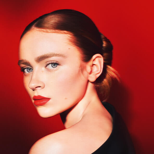 A woman with fair skin and blue eyes gazes over her shoulder against a bold red background, showcasing chic makeup and a stylish hairstyle.