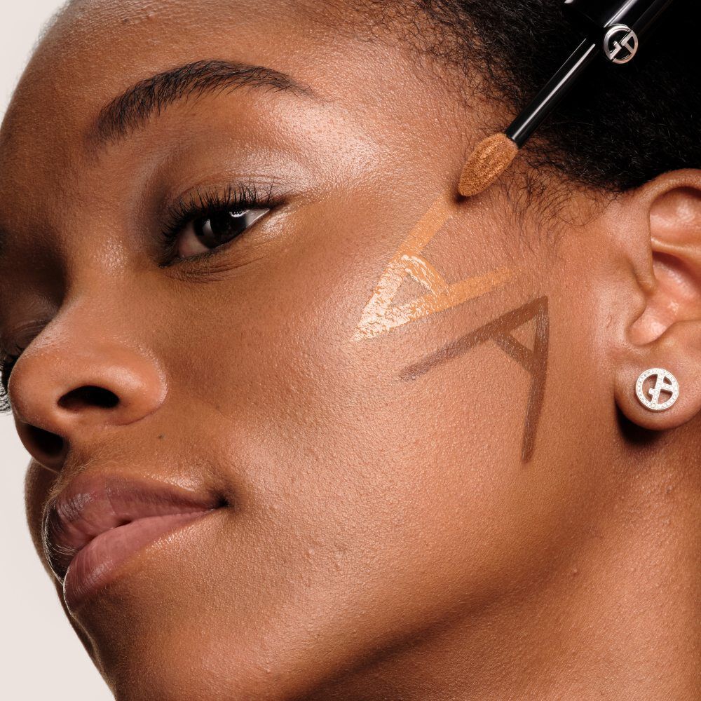 Close-up of a model with deep skin having Luminous Silk Concealer applied to her cheek in a geometric line pattern using the wand applicator.