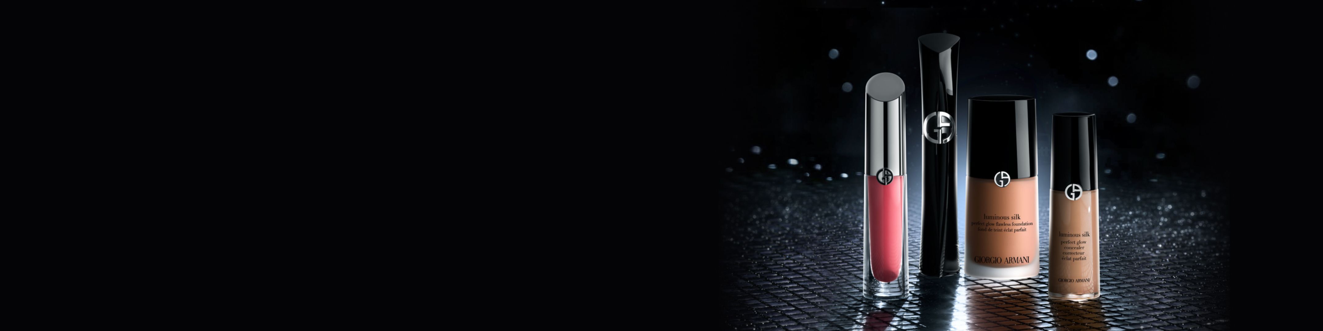 Armani Beauty makeup on dark background: red lip lacquer, black mascara, Luminous Silk foundation and concealer on a reflective surface.