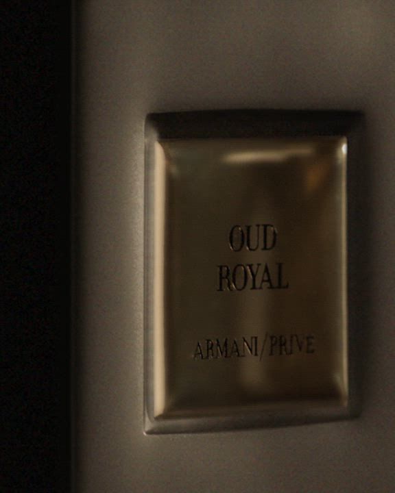 A dark "Armani/Prive OUD ROYAL" fragrance bottle with a black, reflective cap. Overlays of dark, textured wood and a shimmering black flower emphasize the rich, mysterious scent.