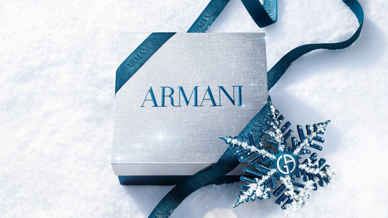 Armani beauty gift box — silver gift box wrapped with blue satin ribbon and a metallic snowflake ornament, resting on fresh white snow.