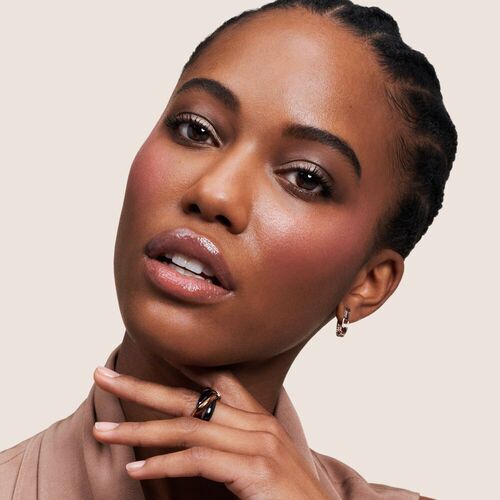 A close-up of a woman with braided hair and makeup, showcasing jewelry and a natural beauty look against a neutral background.