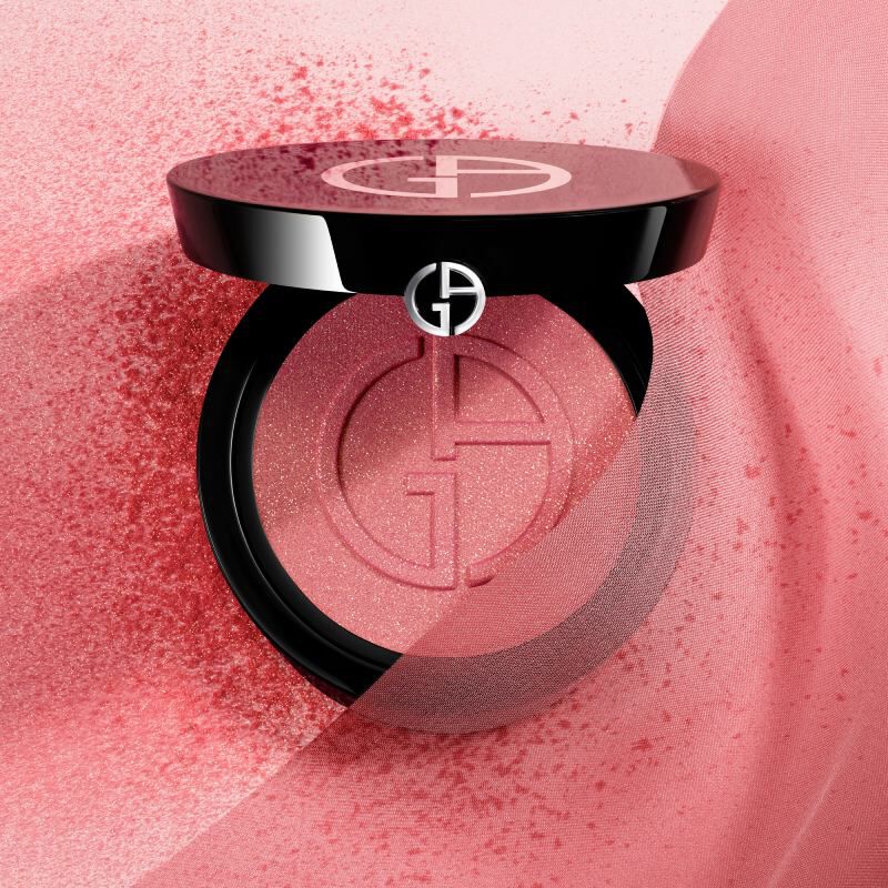 A close-up of a stylish compact of blush, featuring a shiny black lid with a logo, surrounded by soft pink powder and a textured background.