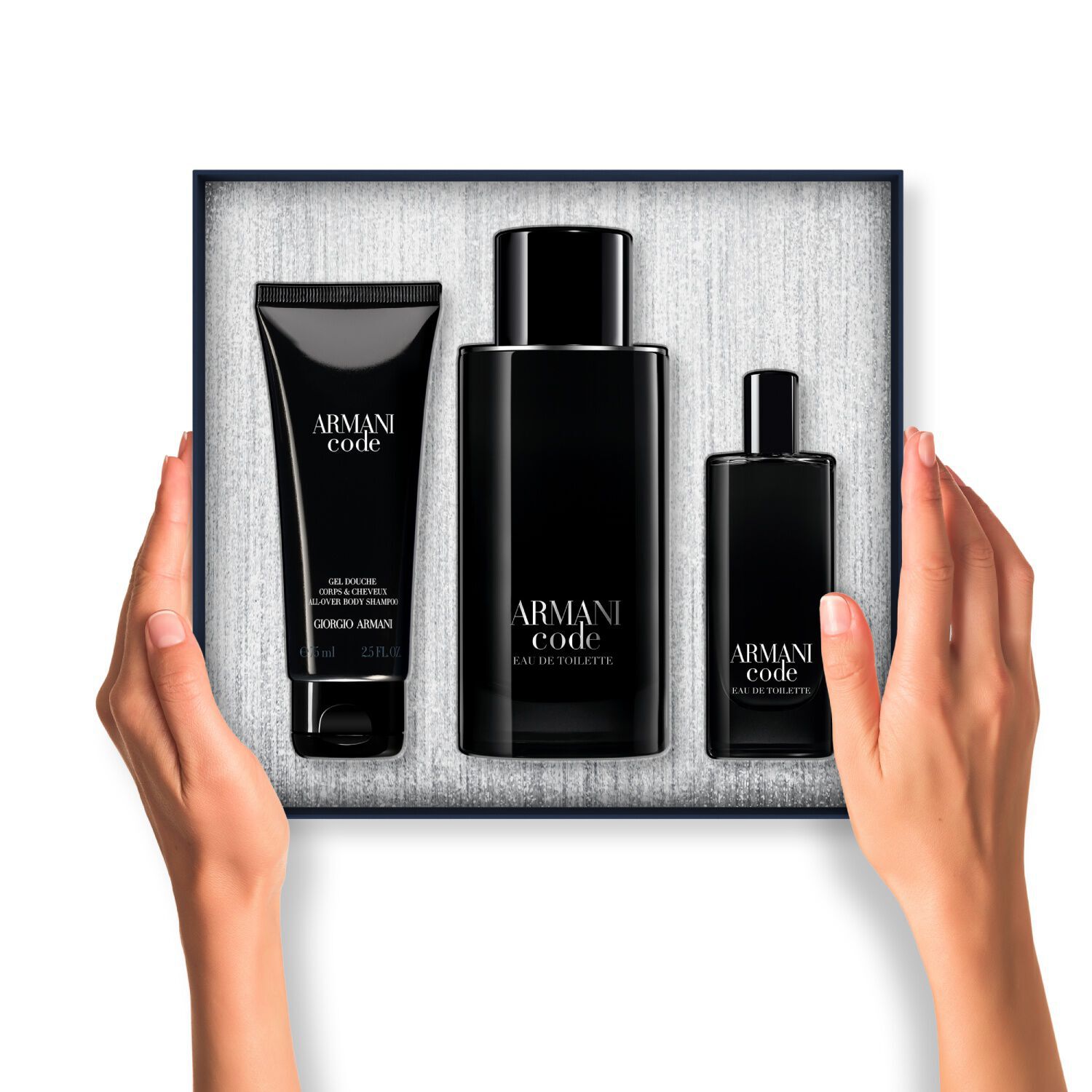 A pair of hands holds an open Armani Code gift box. Inside, three black-packaged products (body gel, large Eau de Toilette, and travel-size Eau de Toilette) are neatly arranged against a textured silver background.