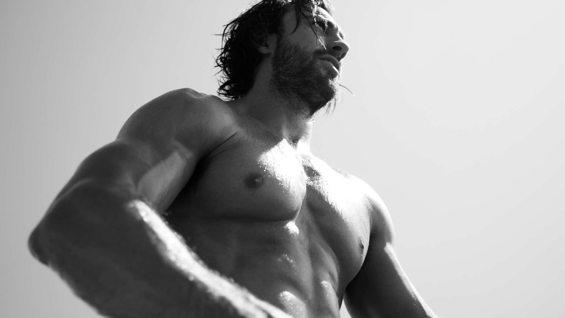 A close-up of a muscular man with wet skin, looking upward against a bright, sunlit background, in black and white.